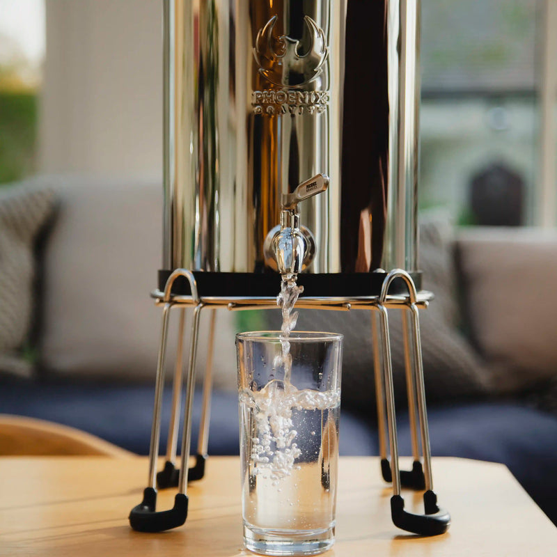 Silver water dispenser pouring water into a glass on a wooden table with a blurred indoor background.